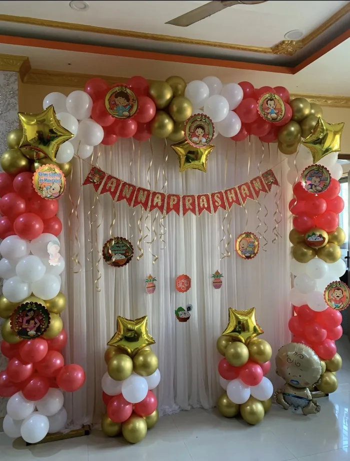 Red, gold and white balloon arch over a drape backdrop with an Annaprashan banner and cartoon cutouts