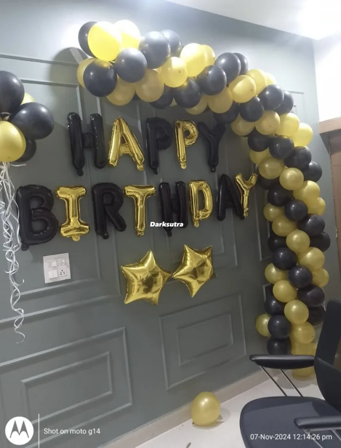 Black and gold balloon arch on a grey panelled wall with foil HAPPY BIRTHDAY letters and gold star balloons