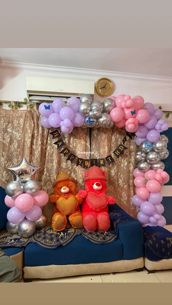 Pastel lilac, pink and silver balloon arch with a Happy Birthday banner and two teddy bears on a blue sofa
