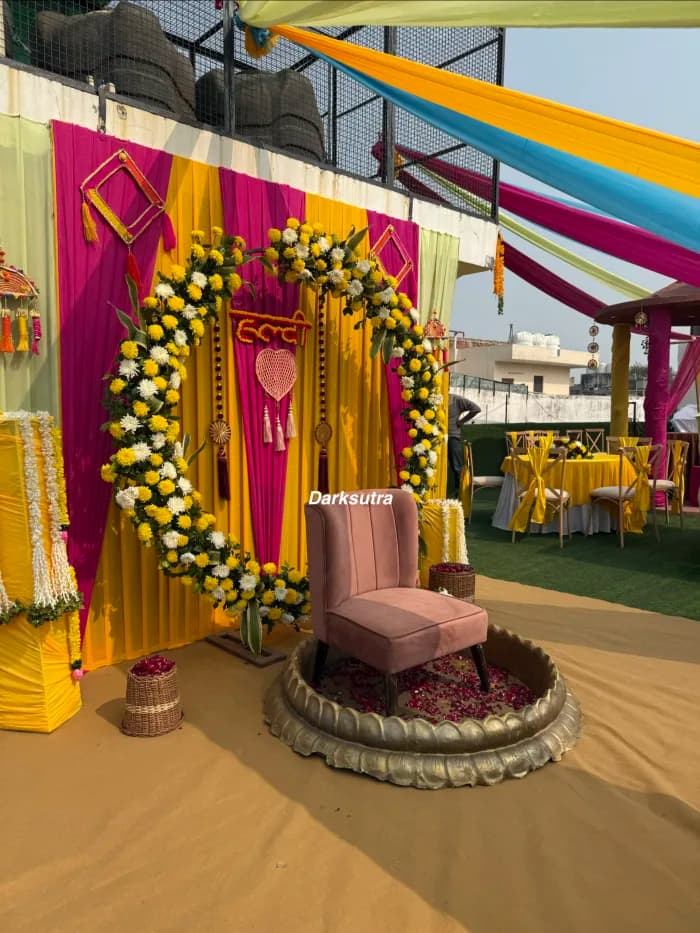 Rooftop haldi setup in pink and yellow drapes with marigold garlands and a cushioned chair on a brass platform