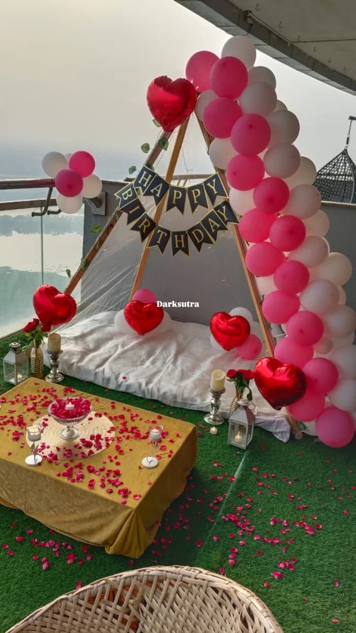 Rooftop teepee date setup with a pink and white balloon arch, red heart balloons, rose petals and a candlelit low table