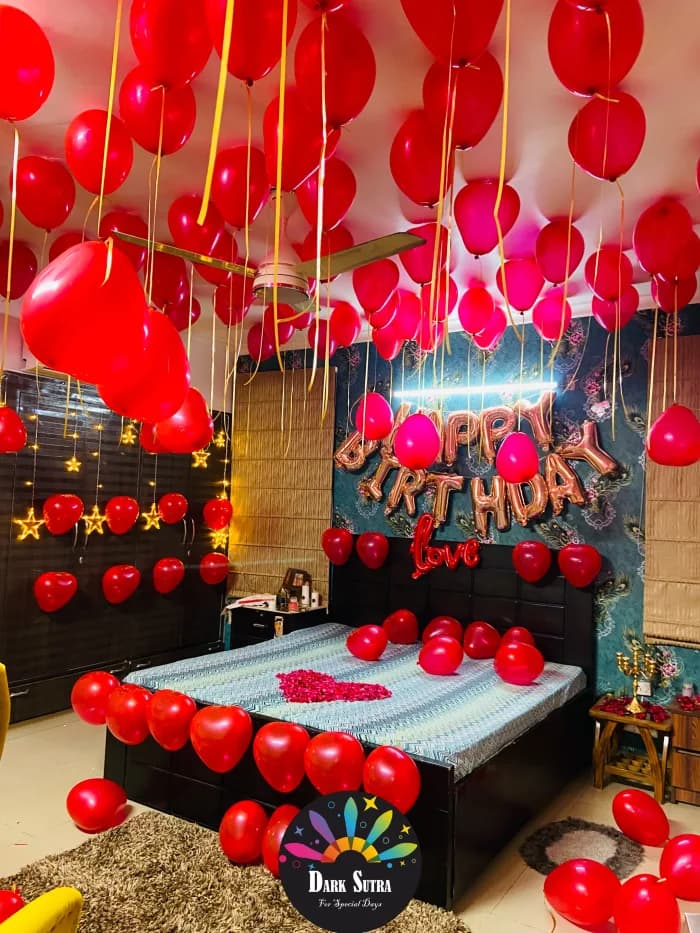 Red heart balloon ceiling with rose gold HAPPY BIRTHDAY letters, a neon love sign and a rose petal heart on the bed