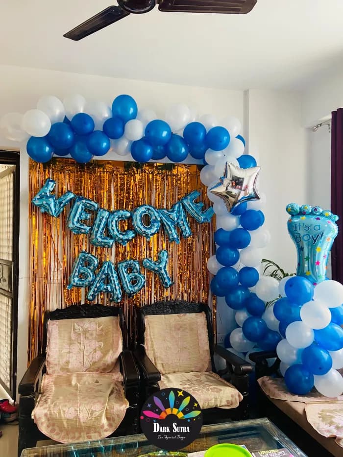 Blue and white balloon arch over a gold foil curtain with WELCOME BABY foil letters and a baby foot balloon